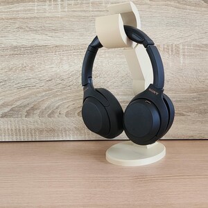 May include: Black over-ear headphones with a gold Sony logo are displayed on a beige stand. The stand is shaped like a stylized ear.