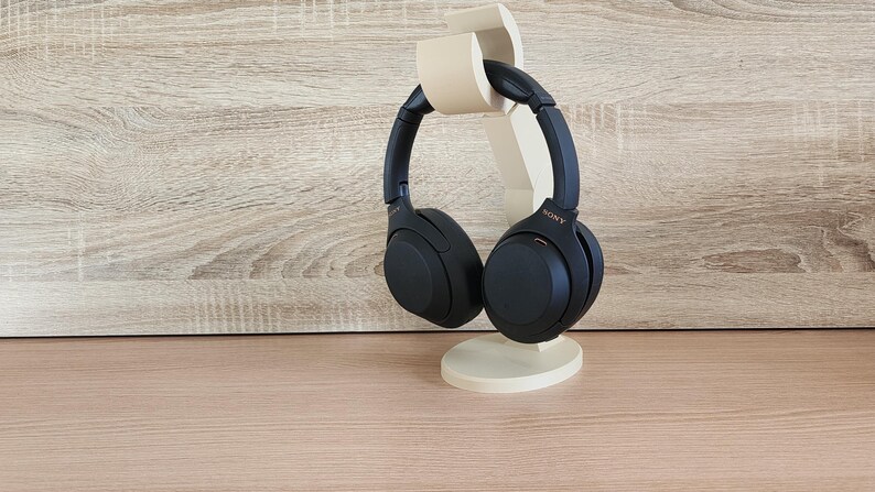 May include: Black over-ear headphones with a gold Sony logo are displayed on a beige stand. The stand is shaped like a stylized ear.