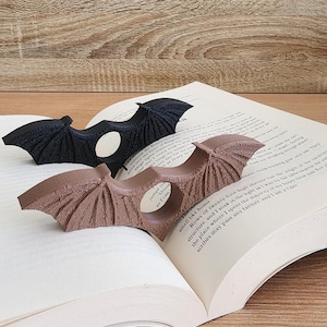 May include: Two bat-shaped bookends, one black and one brown, are holding open a book. The bookends are made of a textured material and have a hole in the center for holding the pages.