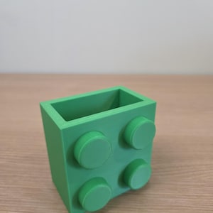 Peut inclure: Un r&eacute;cipient rectangulaire vert vif, ressemblant &agrave; une grosse brique de construction. Le r&eacute;cipient comporte quatre tenons circulaires en relief sur un c&ocirc;t&eacute;. L'objet est fait d'un mat&eacute;riau solide et opaque et repose sur une surface en bois clair.