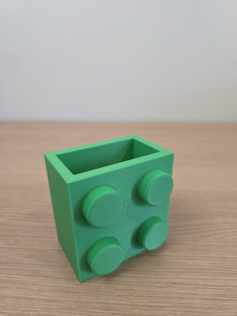 Peut inclure: Un r&eacute;cipient rectangulaire vert vif, ressemblant &agrave; une grosse brique de construction. Le r&eacute;cipient comporte quatre tenons circulaires en relief sur un c&ocirc;t&eacute;. L'objet est fait d'un mat&eacute;riau solide et opaque et repose sur une surface en bois clair.