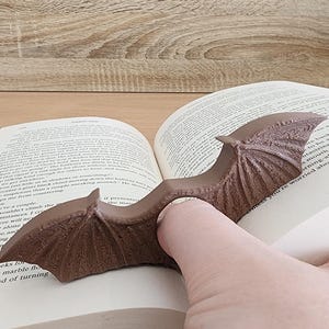 May include: A brown, dragon-shaped book holder with wings spread open, holding a book open. The book holder is made of a textured material and has a realistic design.
