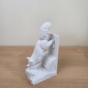 Op de afbeelding: Een lichtgrijze sculptuur van een vos die op een boomstam en een rechthoekige structuur klimt. De vos is gedetailleerd met getextureerde vacht en is gepositioneerd alsof hij actief klimt. De sculptuur staat op een houten oppervlak.