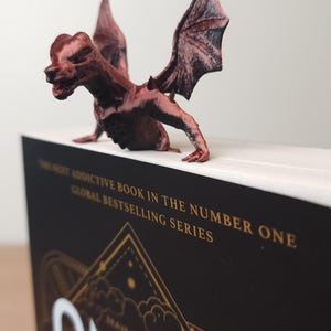 May include: A brown dragon figurine perched on a book. The book has a black cover with gold lettering that reads "ONYX" and "BRAVE THE DARK". The dragon has detailed wings and a menacing expression. The book is part of a global bestselling series.