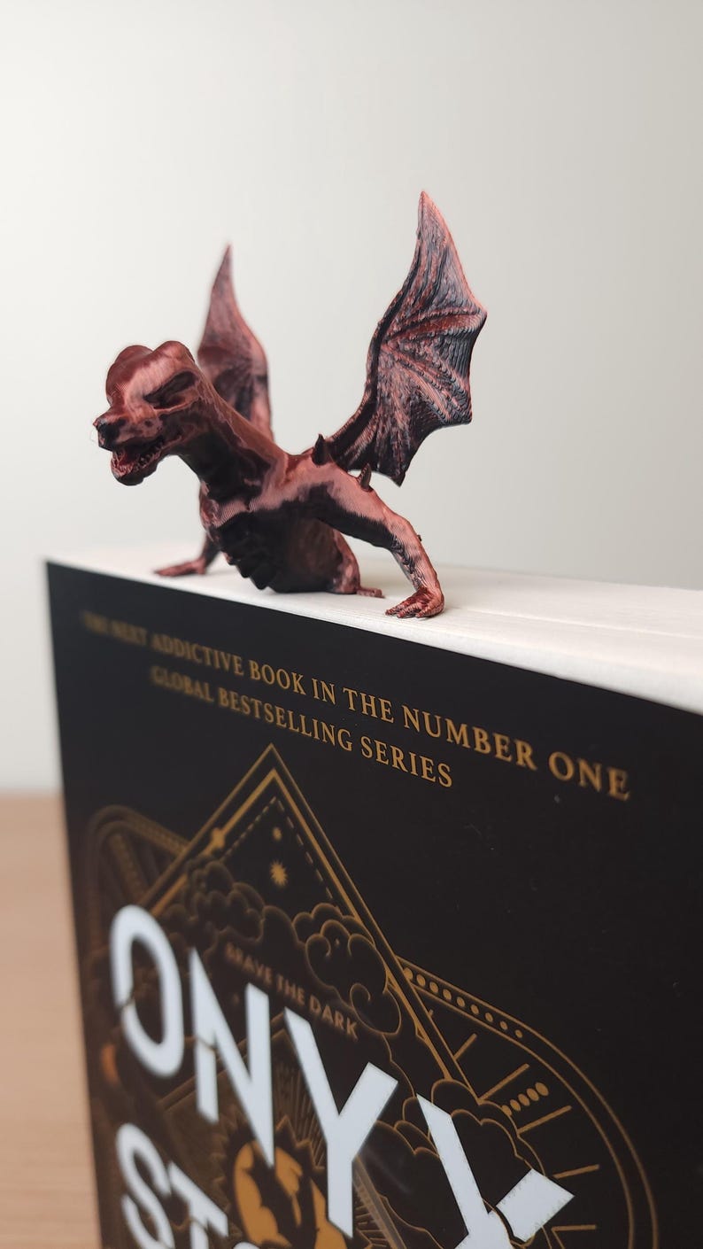 May include: A brown dragon figurine perched on a book. The book has a black cover with gold lettering that reads "ONYX" and "BRAVE THE DARK". The dragon has detailed wings and a menacing expression. The book is part of a global bestselling series.