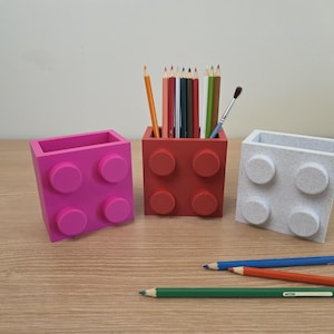Peut inclure: Trois porte-crayons color&eacute;s en forme de briques de jouets. Un rose, un rouge et un blanc. Le porte-crayon rouge contient des crayons et un pinceau. Des crayons sont &eacute;parpill&eacute;s sur la surface en bois.