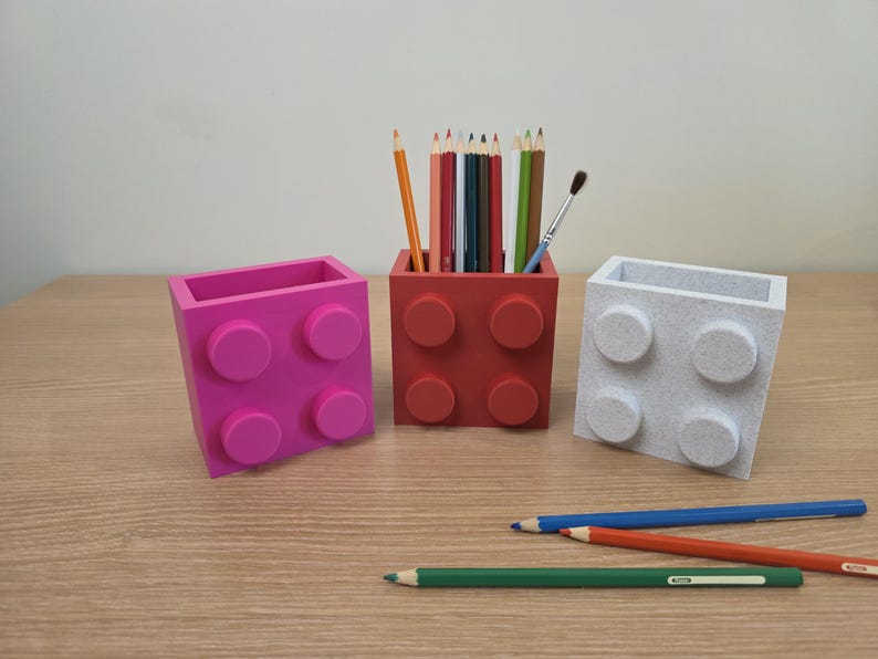 Peut inclure: Trois porte-crayons color&eacute;s en forme de briques de jouets. Un rose, un rouge et un blanc. Le porte-crayon rouge contient des crayons et un pinceau. Des crayons sont &eacute;parpill&eacute;s sur la surface en bois.