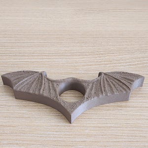 May include: A brown, 3D printed bat-shaped ring holder with a textured surface. The ring holder has a hole in the center for holding a ring.