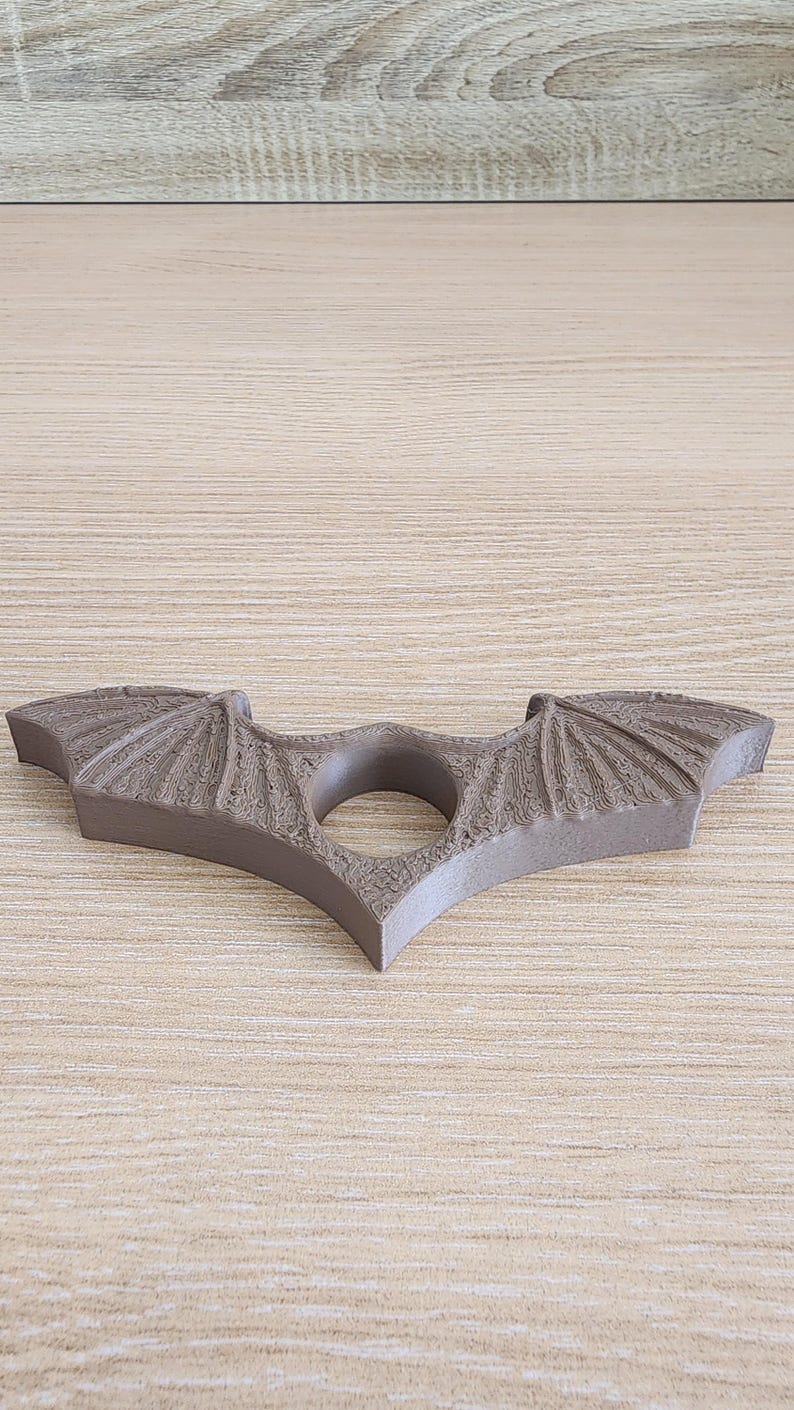 May include: A brown, 3D printed bat-shaped ring holder with a textured surface. The ring holder has a hole in the center for holding a ring.