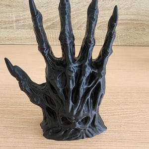 Peut inclure: Une sculpture de main noire stylis&eacute;e avec de longs ongles pointus. La main a une apparence textur&eacute;e et organique, avec des veines visibles et une surface rugueuse. Les doigts sont &eacute;cart&eacute;s, et la sculpture est pos&eacute;e sur une surface en bois.