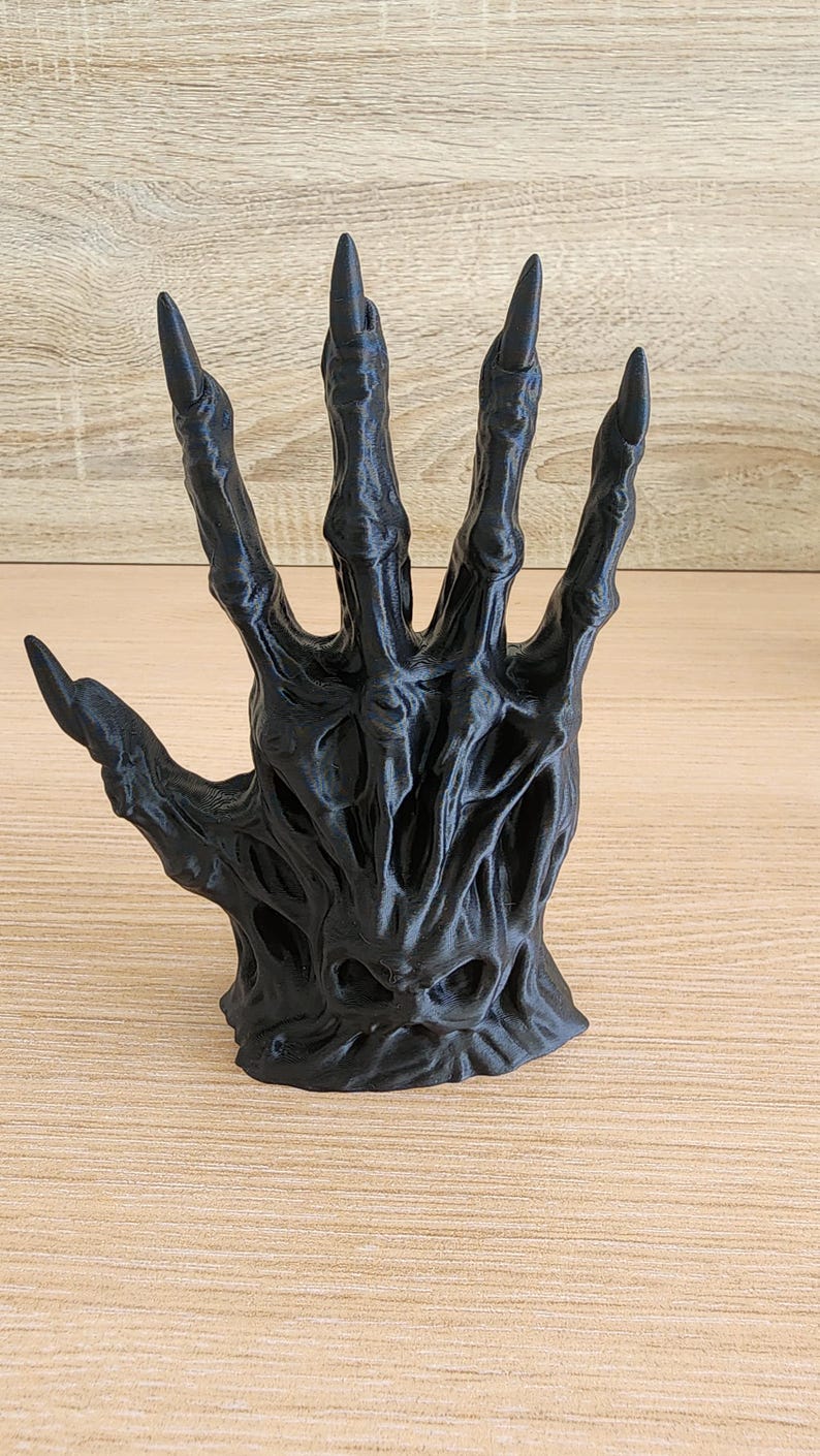 Peut inclure: Une sculpture de main noire stylis&eacute;e avec de longs ongles pointus. La main a une apparence textur&eacute;e et organique, avec des veines visibles et une surface rugueuse. Les doigts sont &eacute;cart&eacute;s, et la sculpture est pos&eacute;e sur une surface en bois.