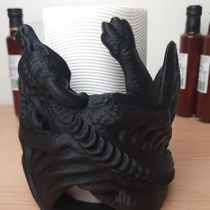 May include: Black 3D printed dragon paper towel holder. The dragon is curled around the paper towel roll and has a detailed design.