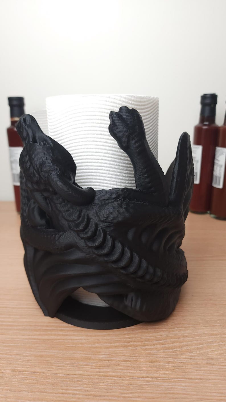 May include: Black 3D printed dragon paper towel holder. The dragon is curled around the paper towel roll and has a detailed design.