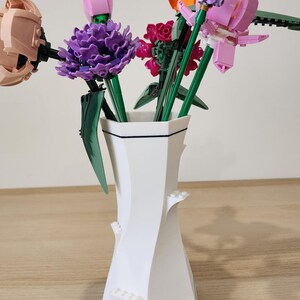 May include: A white ceramic vase with a black trim holds a bouquet of colorful Lego flowers. The flowers include pink, purple, orange, and green.