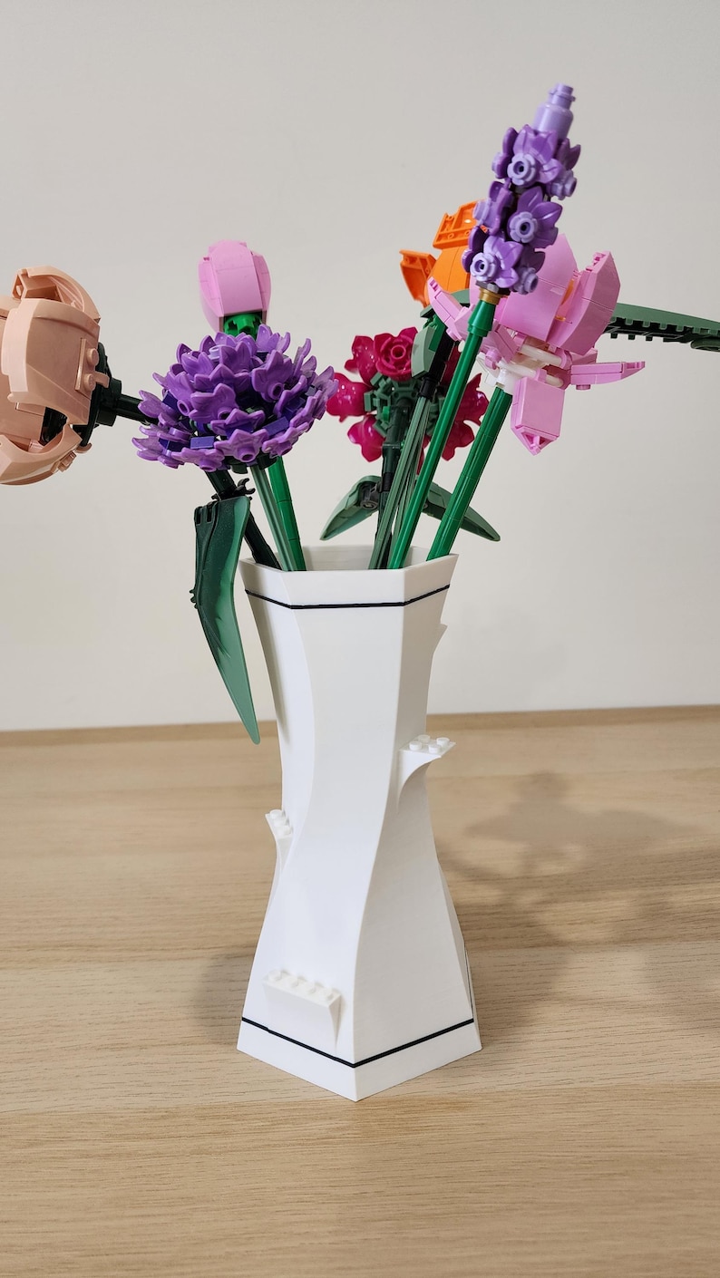 May include: A white ceramic vase with a black trim holds a bouquet of colorful Lego flowers. The flowers include pink, purple, orange, and green.