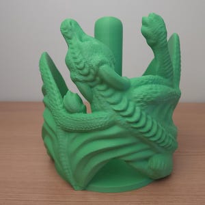 May include: Green 3D printed dragon paper towel holder. The dragon is curled around the holder, with its head and tail visible. The holder is designed to hold a standard roll of paper towels.