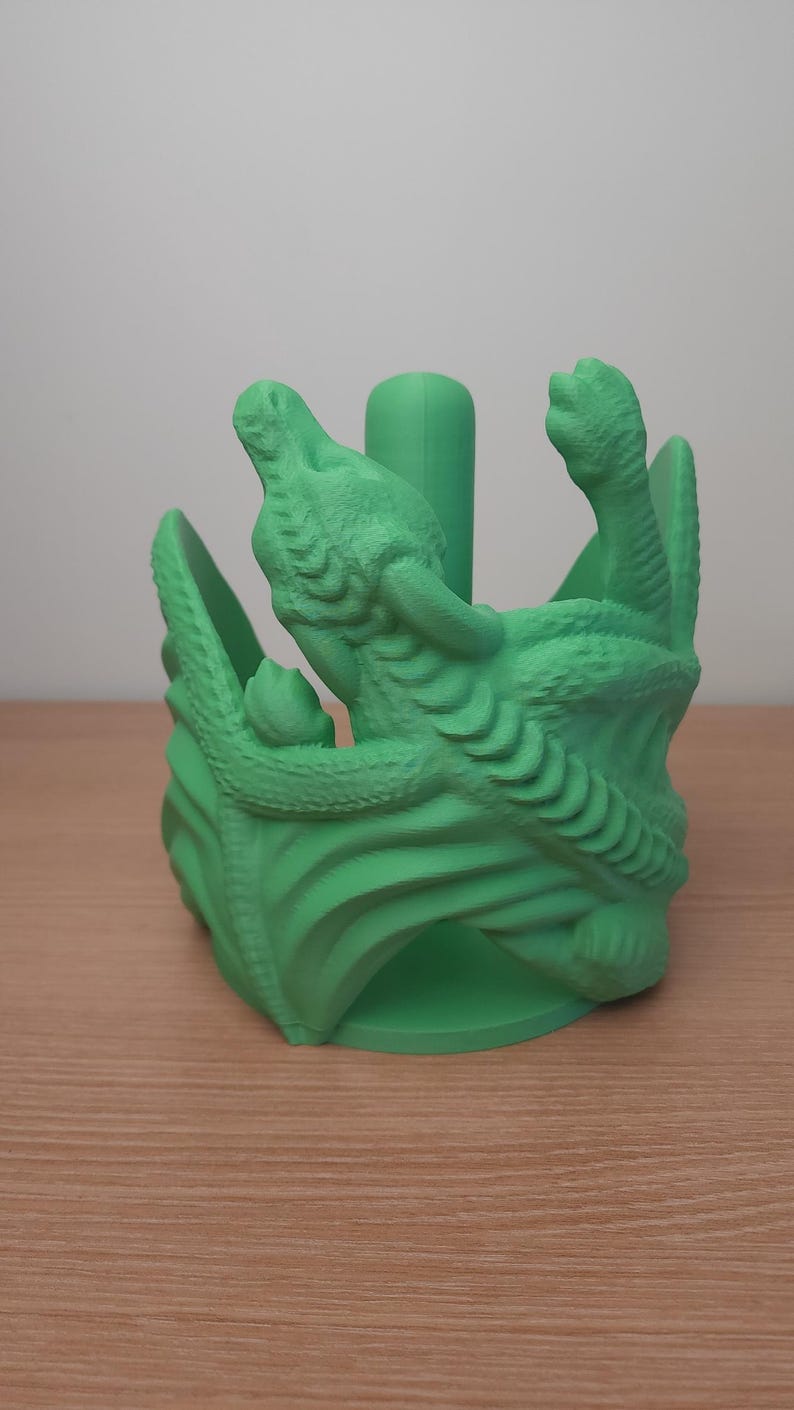 May include: Green 3D printed dragon paper towel holder. The dragon is curled around the holder, with its head and tail visible. The holder is designed to hold a standard roll of paper towels.