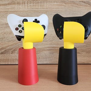 May include: Two video game controllers, one white and one black, are displayed on a yellow and black stand. The stands are shaped like a bird's head with a yellow beak.