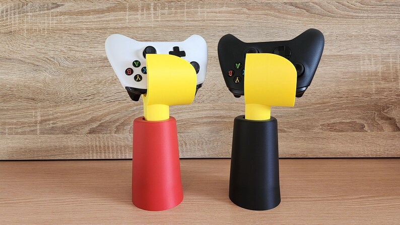 May include: Two video game controllers, one white and one black, are displayed on a yellow and black stand. The stands are shaped like a bird's head with a yellow beak.