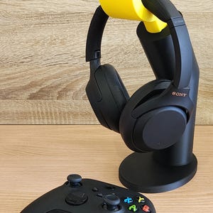 May include: A black gaming headset with yellow earcup holders is on a black stand. A black Xbox controller is on a wooden surface.