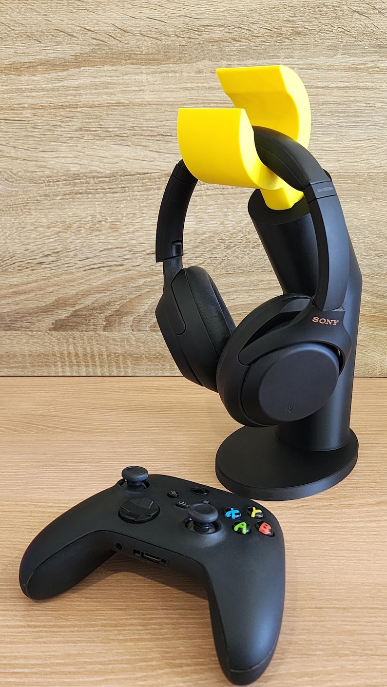 May include: A black gaming headset with yellow earcup holders is on a black stand. A black Xbox controller is on a wooden surface.