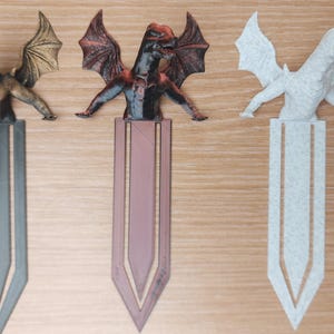 May include: Three dragon-themed bookmarks. The first is bronze, the second is red and black, and the third is white. Each dragon is perched atop a bookmark with a pointed end. The bookmarks are on a wooden surface.