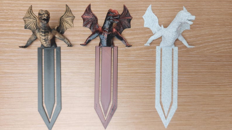 May include: Three dragon-themed bookmarks. The first is bronze, the second is red and black, and the third is white. Each dragon is perched atop a bookmark with a pointed end. The bookmarks are on a wooden surface.