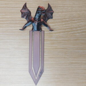 May include: A dragon-themed bookmark with a detailed, multi-colored dragon figure atop a lavender-colored bookmark. The dragon has outstretched wings and a detailed body. The bookmark is made of a durable material.