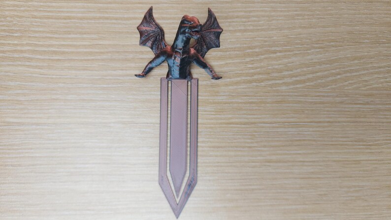 May include: A dragon-themed bookmark with a detailed, multi-colored dragon figure atop a lavender-colored bookmark. The dragon has outstretched wings and a detailed body. The bookmark is made of a durable material.