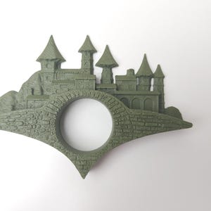 May include: A green 3D printed bookmark shaped like a castle with a hole in the middle for a page. The castle has multiple towers and is set on a hill.