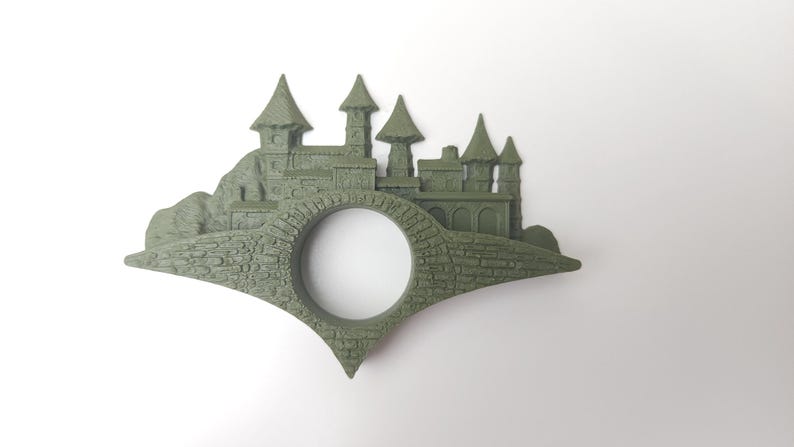 May include: A green 3D printed bookmark shaped like a castle with a hole in the middle for a page. The castle has multiple towers and is set on a hill.