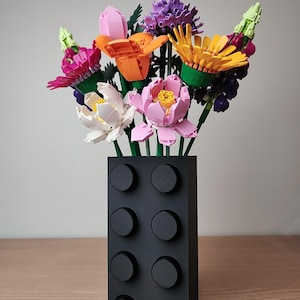 K&ouml;nnte beinhalten: Eine schwarze Vase in Form eines Bausteins h&auml;lt einen bunten Blumenstrau&szlig;. Die Blumen bestehen aus verschiedenfarbigen Kunststoffteilen, darunter Rosa, Orange, Gelb und Lila. Die Vase hat acht kreisf&ouml;rmige Noppen auf der Vorderseite.