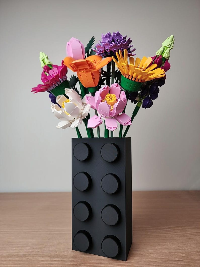 K&ouml;nnte beinhalten: Eine schwarze Vase in Form eines Bausteins h&auml;lt einen bunten Blumenstrau&szlig;. Die Blumen bestehen aus verschiedenfarbigen Kunststoffteilen, darunter Rosa, Orange, Gelb und Lila. Die Vase hat acht kreisf&ouml;rmige Noppen auf der Vorderseite.