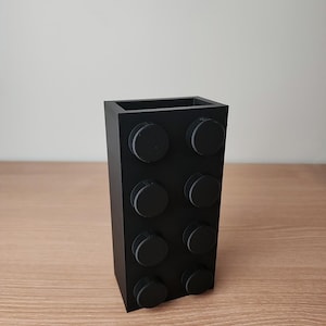 K&ouml;nnte beinhalten: Eine schwarze, rechteckige Vase, die einem gro&szlig;en Baustein nachempfunden ist. Die Vase hat acht kreisf&ouml;rmige Noppen auf der Vorderseite und besteht aus einem mattschwarzen Material. Die Vase steht auf einer Holzoberfl&auml;che.