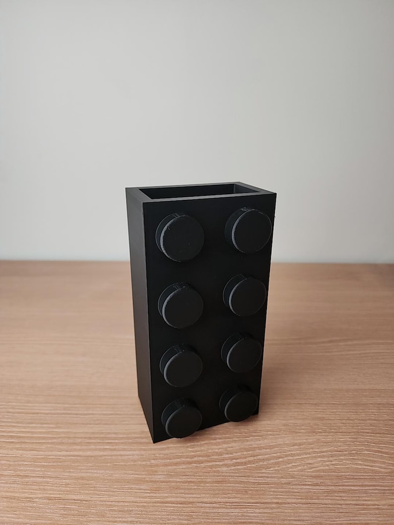 K&ouml;nnte beinhalten: Eine schwarze, rechteckige Vase, die einem gro&szlig;en Baustein nachempfunden ist. Die Vase hat acht kreisf&ouml;rmige Noppen auf der Vorderseite und besteht aus einem mattschwarzen Material. Die Vase steht auf einer Holzoberfl&auml;che.