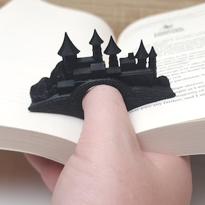 May include: A black 3D printed bookmark shaped like a castle. The bookmark is being used to hold a book open.