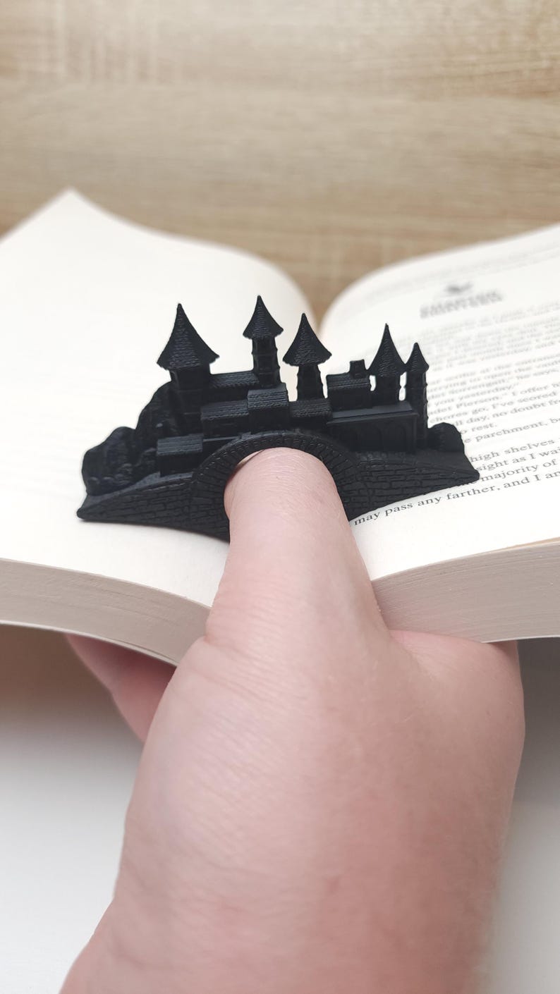 May include: A black 3D printed bookmark shaped like a castle. The bookmark is being used to hold a book open.
