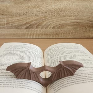 May include: A brown bat-shaped bookmark is placed between the pages of an open book. The book is open to a page with text about a character named Orion. The book is resting on a wooden surface.