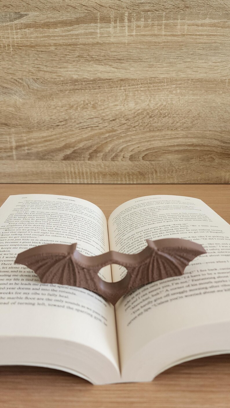 May include: A brown bat-shaped bookmark is placed between the pages of an open book. The book is open to a page with text about a character named Orion. The book is resting on a wooden surface.