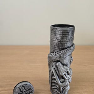 May include: A cylindrical, gray container with a textured, brick-like pattern. A detailed dragon wraps around the container. A separate, circular lid with a geometric design sits nearby. The container is likely a decorative item.