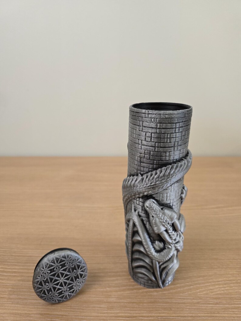 May include: A cylindrical, gray container with a textured, brick-like pattern. A detailed dragon wraps around the container. A separate, circular lid with a geometric design sits nearby. The container is likely a decorative item.