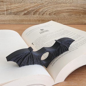 May include: A black bat-shaped bookmark with a small silver charm that says "well" and "I'd" on it. The bookmark is placed in a book with the text "the place where I spent the time" and "scribes may pass any further and so" visible.