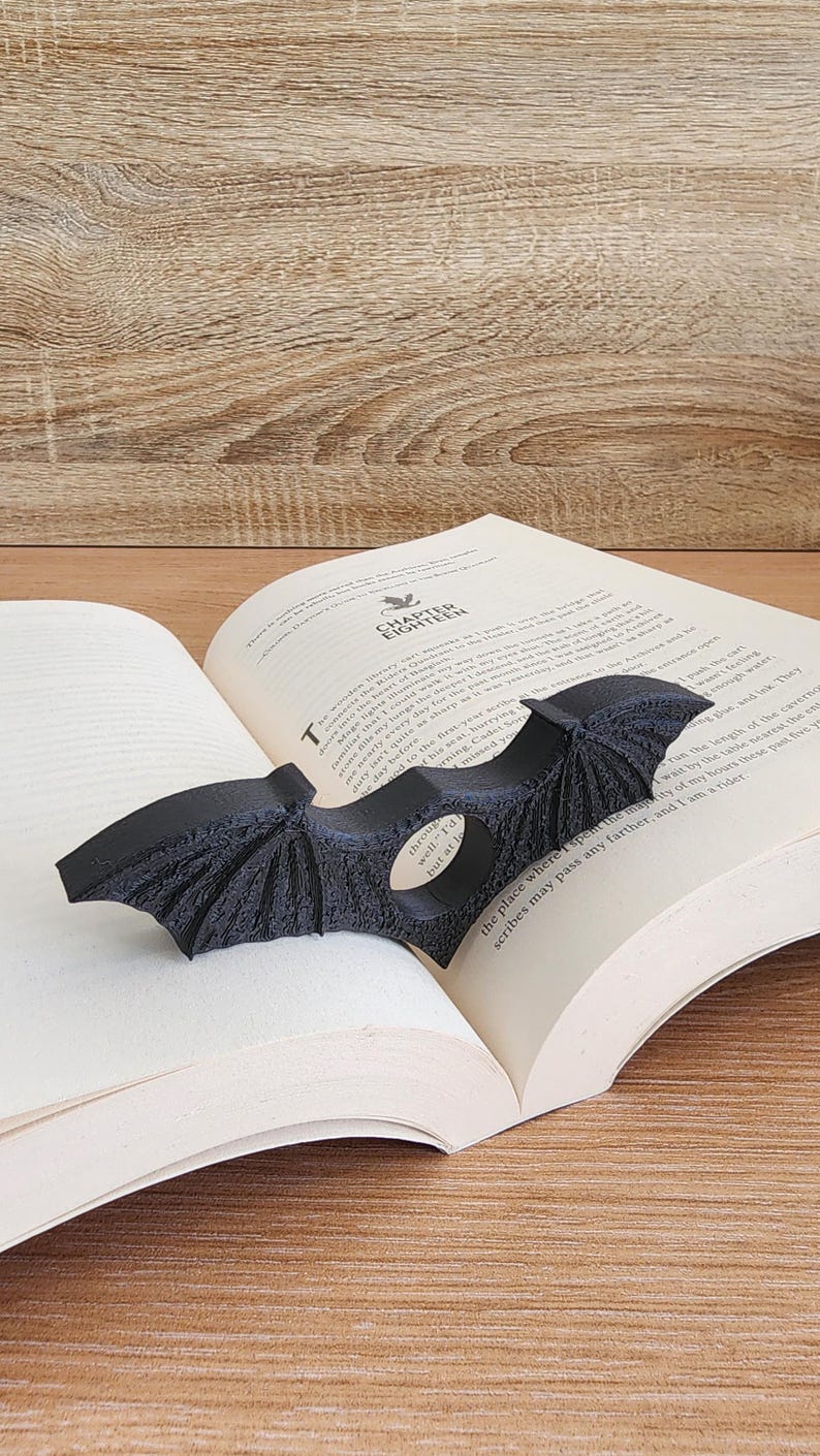 May include: A black bat-shaped bookmark with a small silver charm that says "well" and "I'd" on it. The bookmark is placed in a book with the text "the place where I spent the time" and "scribes may pass any further and so" visible.