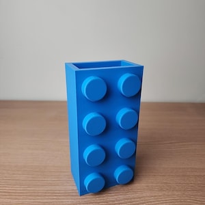 K&ouml;nnte beinhalten: Eine leuchtend blaue Vase in Form eines gro&szlig;en Bausteins mit acht kreisf&ouml;rmigen Noppen. Die rechteckige Vase besteht aus einem festen Material und steht auf einer Holzoberfl&auml;che. Die Vase ist etwa 15 cm hoch.