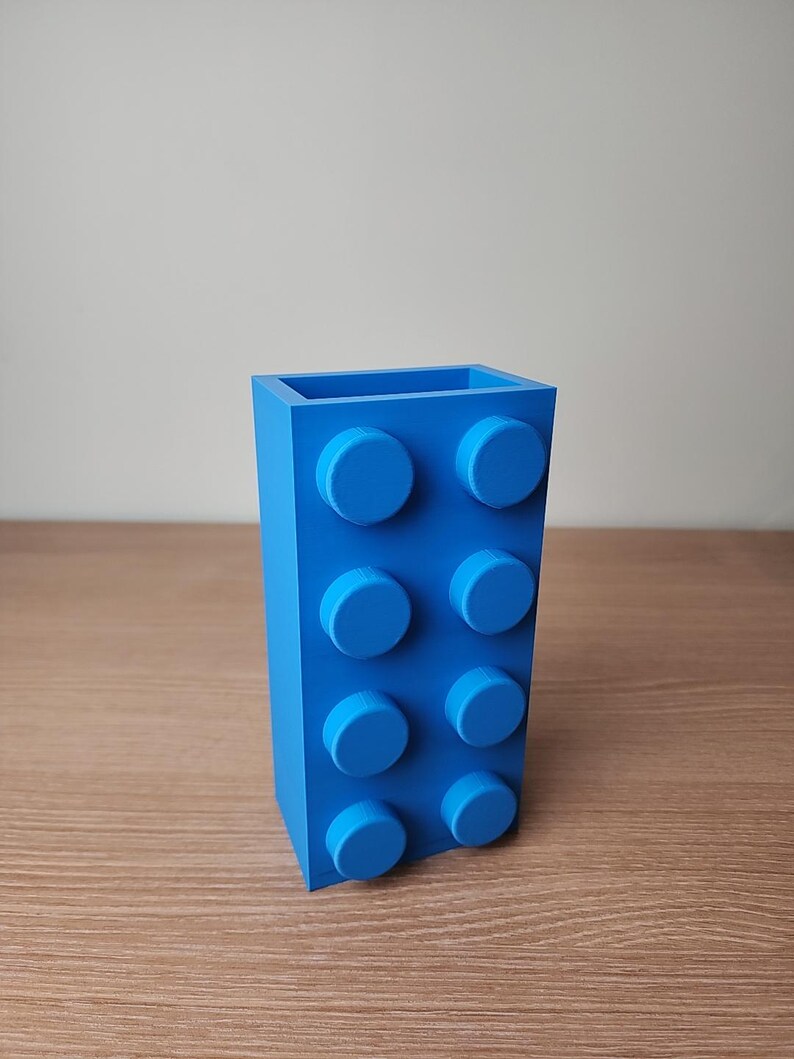 K&ouml;nnte beinhalten: Eine leuchtend blaue Vase in Form eines gro&szlig;en Bausteins mit acht kreisf&ouml;rmigen Noppen. Die rechteckige Vase besteht aus einem festen Material und steht auf einer Holzoberfl&auml;che. Die Vase ist etwa 15 cm hoch.