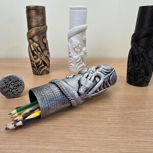 May include: A collection of decorative pencil holders and a small round object. The pencil holders are cylindrical with a dragon and snake design. One is silver, one is bronze, one is white, and one is black. Pencils are visible in the silver holder.