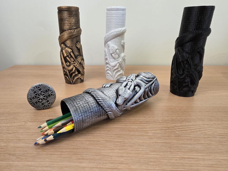 May include: A collection of decorative pencil holders and a small round object. The pencil holders are cylindrical with a dragon and snake design. One is silver, one is bronze, one is white, and one is black. Pencils are visible in the silver holder.