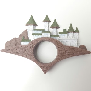 May include: A brown and white ceramic ring holder shaped like a bridge with a small castle on top. The castle has green roofs and white walls.