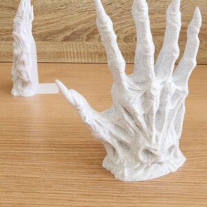 Peut inclure: Une sculpture blanche et textur&eacute;e d'une main squelettique aux doigts allong&eacute;s. La main est ouverte, la paume vers le haut. Une sculpture plus petite et similaire est derri&egrave;re. Les sculptures sont sur une surface en bois.