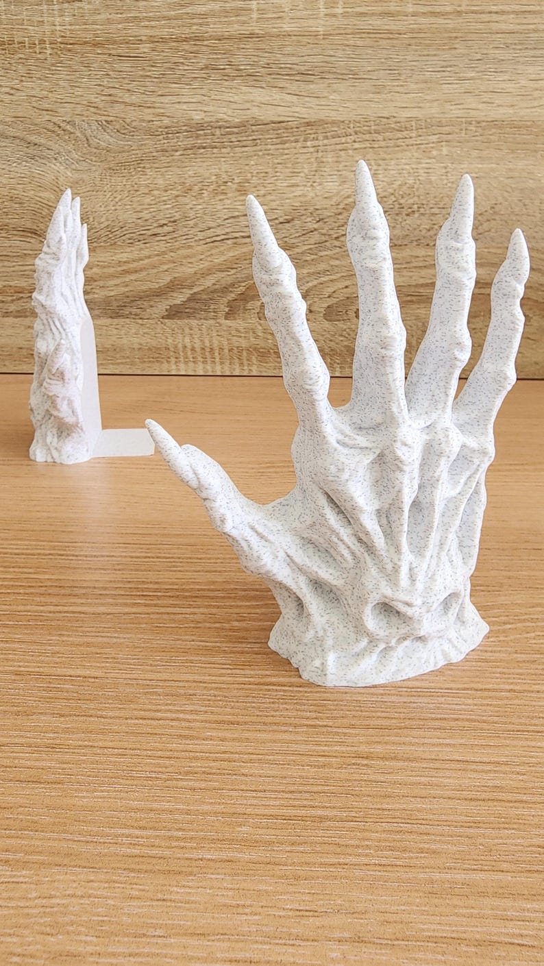 Peut inclure: Une sculpture blanche et textur&eacute;e d'une main squelettique aux doigts allong&eacute;s. La main est ouverte, la paume vers le haut. Une sculpture plus petite et similaire est derri&egrave;re. Les sculptures sont sur une surface en bois.